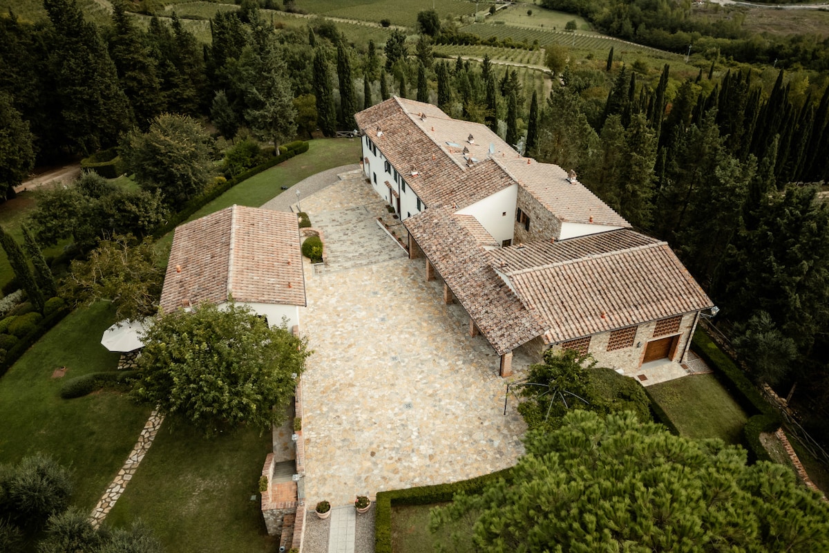 An aerial view captures the expansive grounds of a Tuscan estate, showcasing a rustic farmhouse with terracotta roofs. Manicured gardens surround the structure, with cypress trees lining the perimeter. The stone pathway leads towards a serene outdoor space, emphasizing the peaceful countryside setting.