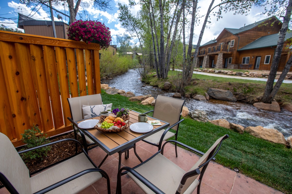 A stream-side patio is shown with a round table and four chairs. A platter of fresh fruit is displayed alongside plates and a cup. Lush greenery and the flowing Fall River can be seen in the background, while nearby buildings feature rustic designs and wood accents.