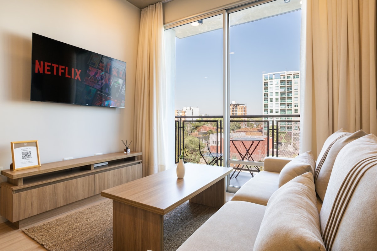A modern living area features a light-colored sofa positioned in front of a wooden coffee table. A flat-screen TV displays the Netflix logo, while large windows provide a view of the city skyline. Sheer curtains allow natural light to brighten the space.