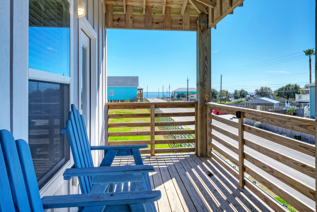 A balcony is shown with two blue chairs positioned for relaxation. The railing frames a view of the surrounding area, including distant structures and palm trees. Bright sunlight enhances the inviting outdoor space, perfect for enjoying the fresh air.