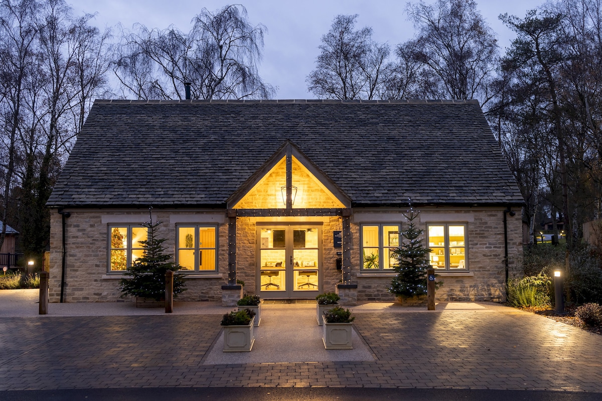 The lodge's exterior is illuminated warmly at dusk, featuring a stone facade and a gabled roof. Symmetrical windows framed with trees provide a welcoming entrance, while evenly spaced ground lighting highlights the pathway and beautifully landscaped surroundings.