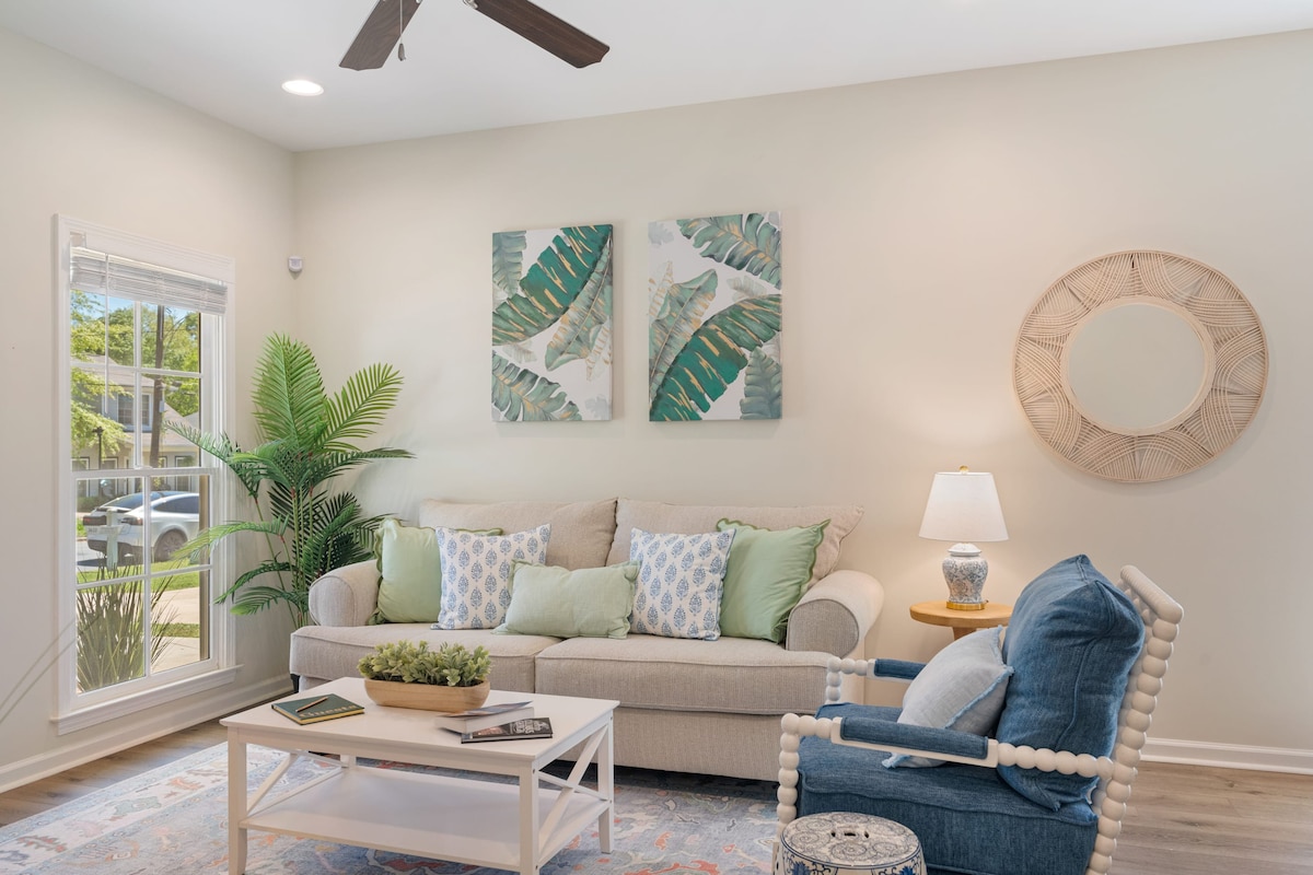 A cozy living room features a beige sofa adorned with decorative pillows and a light blue armchair with unique detailing. Two pieces of wall art showcase green foliage, while a small round table holds a potted plant. Natural light brightens the space through large windows.
