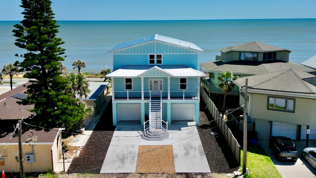A modern, three-story beachfront house is shown, featuring a light blue exterior and a spacious driveway. Palm trees are visible nearby, complemented by a clear ocean view in the background. The entrance is framed by a set of stairs leading to the porch.
