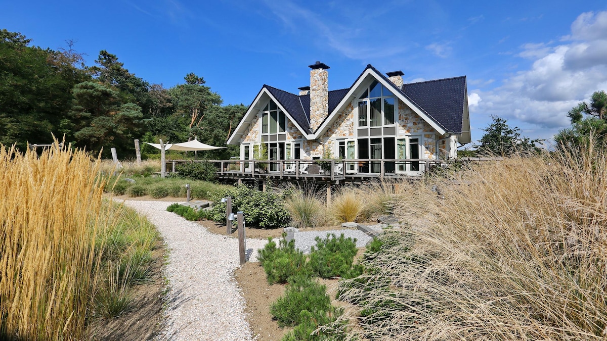 The exterior of the villa displays a striking architectural design featuring large windows and a high-pitched roof. A pathway made of gravel leads through natural landscaping, complemented by tall grasses and flowering plants that frame the entrance, enhancing the serene outdoor environment.