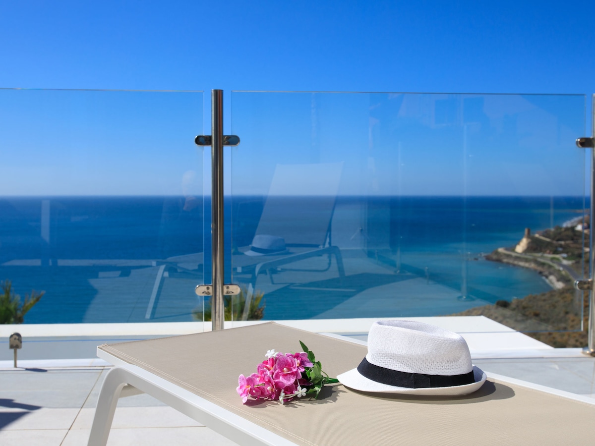 A sun lounger is positioned on a terrace, featuring a light fabric and a flat surface. A white straw hat with a black band rests beside a cluster of pink flowers. In the background, a panoramic view of the sea and coastline is visible under a clear blue sky.