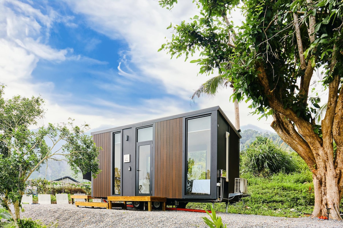 A modern tiny house features a sleek design with large windows, allowing natural light to fill the interior. It is set against a backdrop of lush greenery and tropical trees, surrounded by a gravel pathway that complements the peaceful mountainous environment.