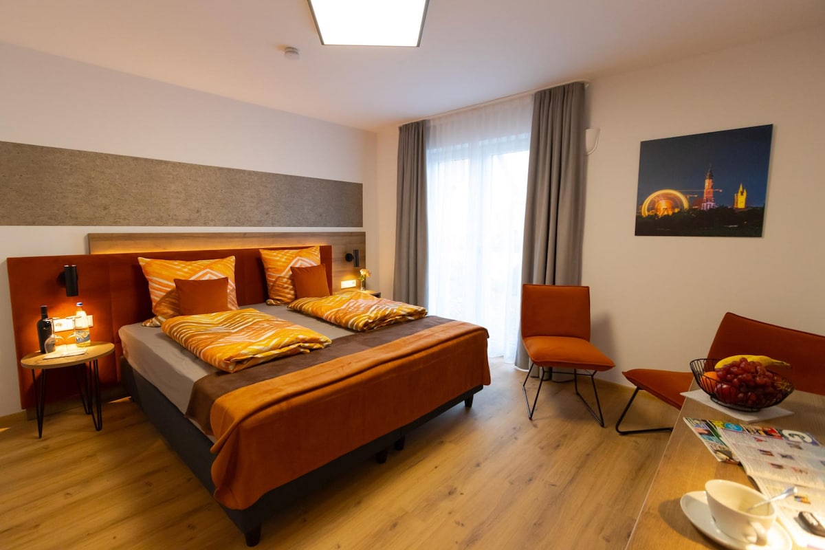 A welcoming guest room features a spacious bed with patterned orange and brown linens. Two warm-toned chairs complement a small table holding fresh fruit. Natural light filters through a large window with sheer curtains, enhancing the warm ambiance of the space.