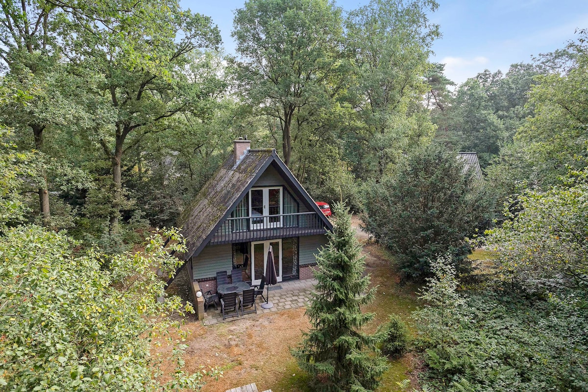 The bungalow is situated in a wooded area, surrounded by trees and shrubs. A patio area with rustic furniture is visible in front of the structure, which features a sloped roof and a balcony on the upper floor. Natural greenery frames the scene.