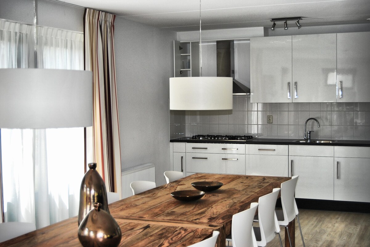 A modern dining area features a large wooden table surrounded by white chairs. Above the table, two pendant lights provide illumination. The kitchen is visible in the background, equipped with sleek cabinetry and appliances, complemented by natural light from large windows draped with striped curtains.