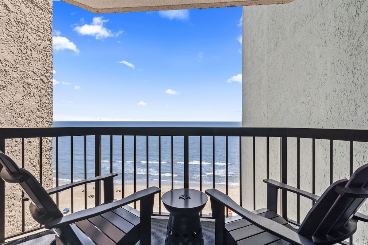 A private balcony offers ocean views, with two black adirondack chairs positioned beside a round side table. The horizon showcases the calm sea and blue sky, while the sandy beach is visible below.