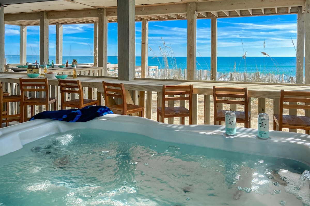 A hot tub is positioned in a shaded covered patio area, offering views of the ocean through large open spaces. The area is furnished with a pub-height table and several chairs, creating a relaxed atmosphere for enjoying cocktails or meals while overlooking the beach.