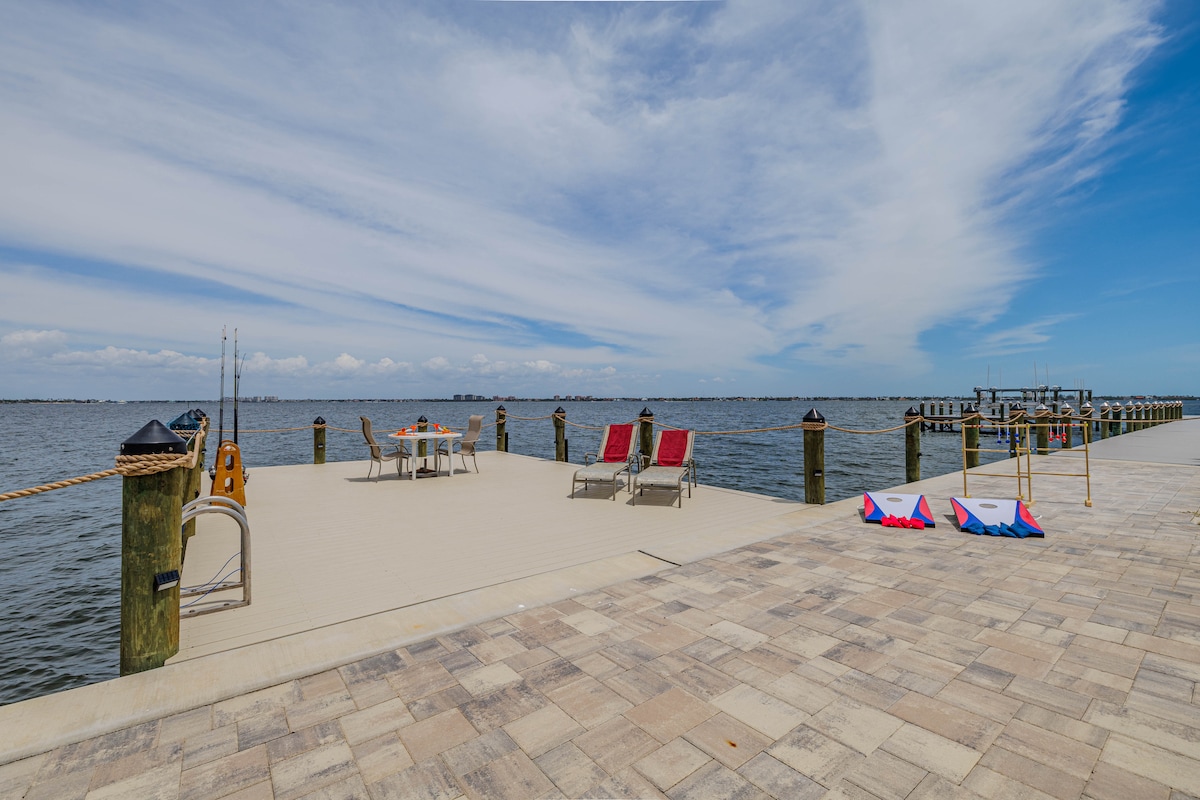 A spacious dock extends into the river, featuring a fishing rod holder and a table set for outdoor dining. Reclining chairs in vibrant colors provide comfort, while sun loungers are positioned beside the water for relaxation. A serene river view stretches into the horizon.