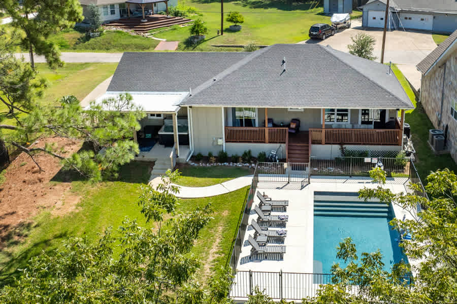 An aerial view of a well-maintained home featuring a spacious covered porch with seating. A rectangular swimming pool is surrounded by lounge chairs, and a neatly landscaped yard adds to the serene atmosphere. Nearby, the driveway and additional outdoor spaces are visible.