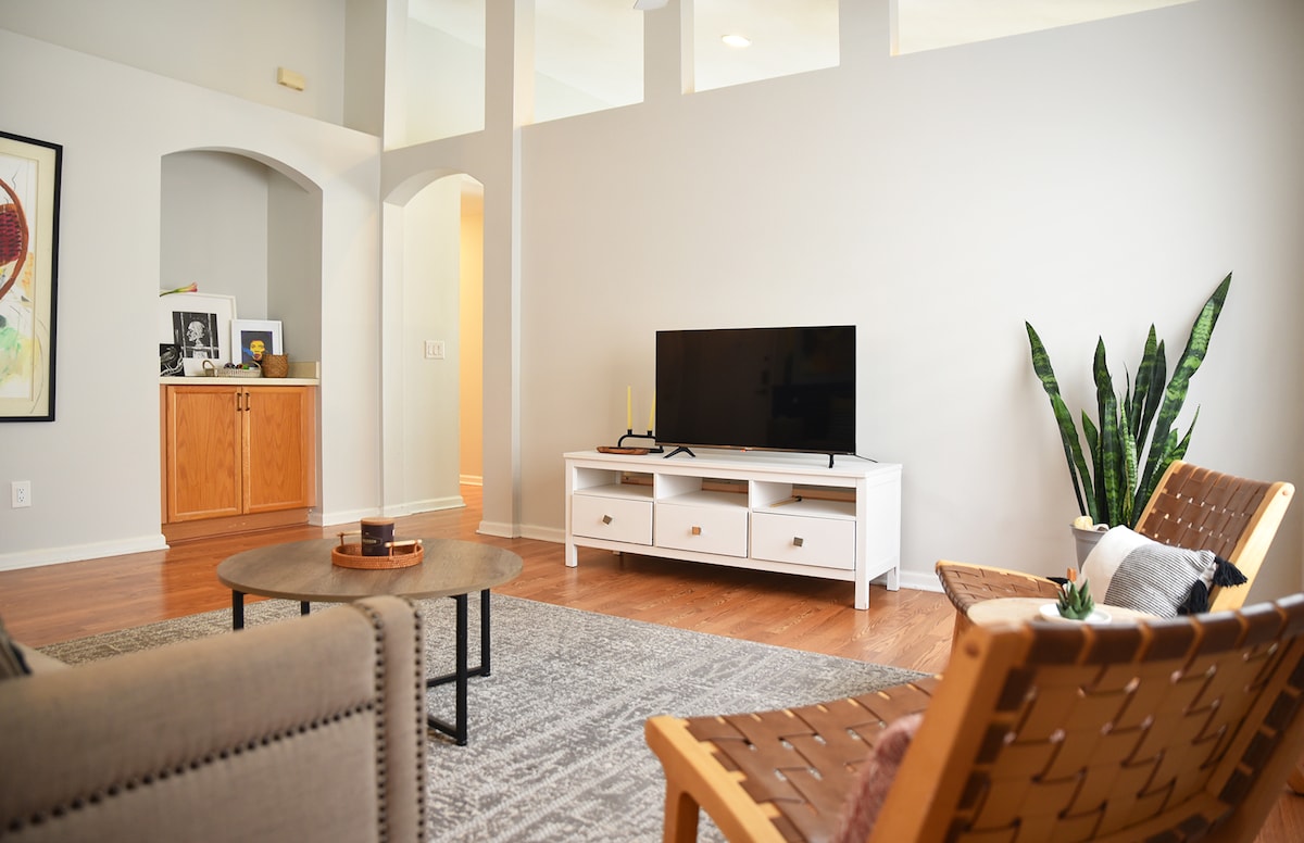 A spacious living area features a large flat-screen TV positioned on a white media console. Two stylish chairs are arranged around a circular coffee table, which is accented by decorative items. A tall plant adds a touch of greenery, complemented by neutral-colored walls and warm wooden flooring.