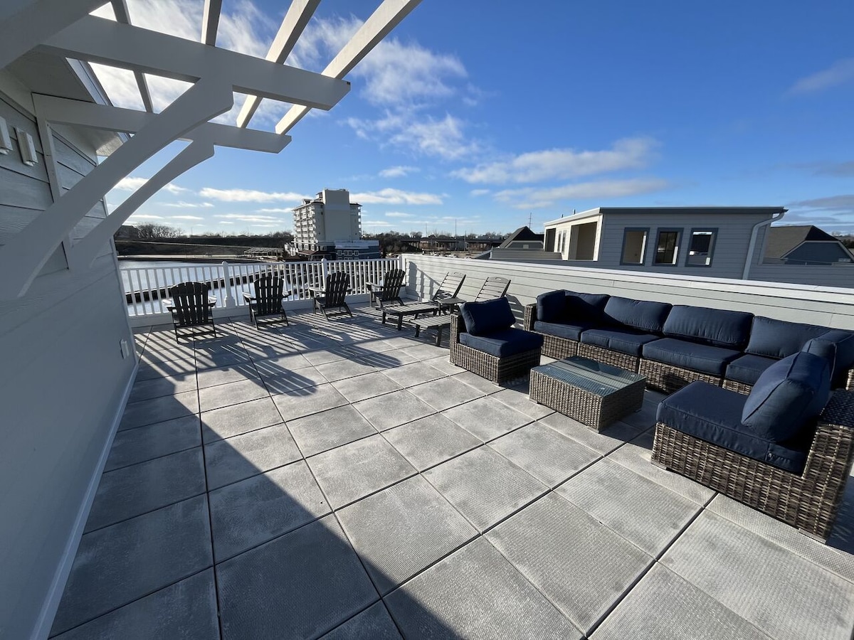 An expansive rooftop patio features ample seating with a sectional sofa and individual chairs arranged around a coffee table. The area is filled with natural light, and clear views of the surrounding landscape can be observed under a bright blue sky.