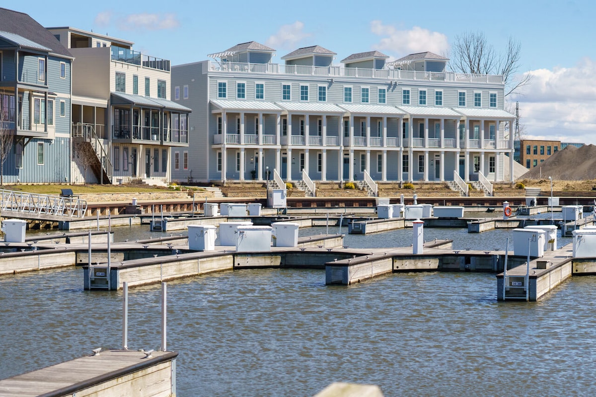 A waterfront view showcases a row of modern townhomes with multiple levels and rooftop patios. The marina features a series of empty boat slips along the calm water, framed by clear skies and a hints of greenery in the background.