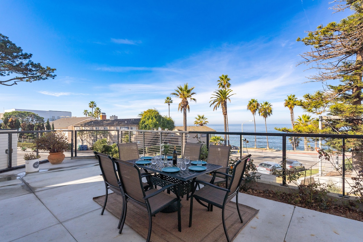 An outdoor patio area features a round dining table surrounded by six chairs, set against a backdrop of ocean views and palm trees. The concrete surface is complemented by a natural fiber rug, while sunlight brightens the inviting atmosphere.