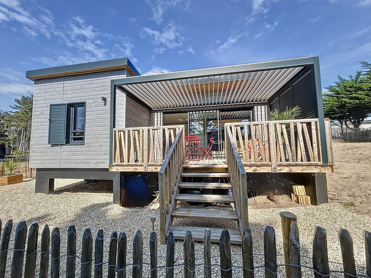 A modern cottage features a raised wooden terrace with a covered area, providing seating for relaxation. The exterior showcases a mix of wood and grey siding, complemented by large sliding glass doors that open to the interior. Surrounding landscaping includes a gravel area and decorative fencing.