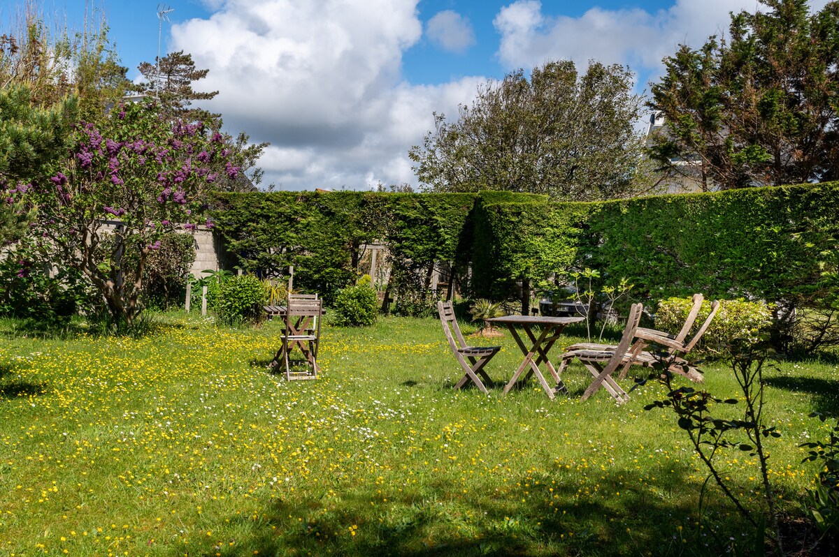 A lush garden area is surrounded by tall hedges and trees, featuring a grassy space dotted with yellow flowers. Wooden outdoor furniture including a table and chairs is positioned for relaxation, complementing the natural setting under a partly cloudy sky.