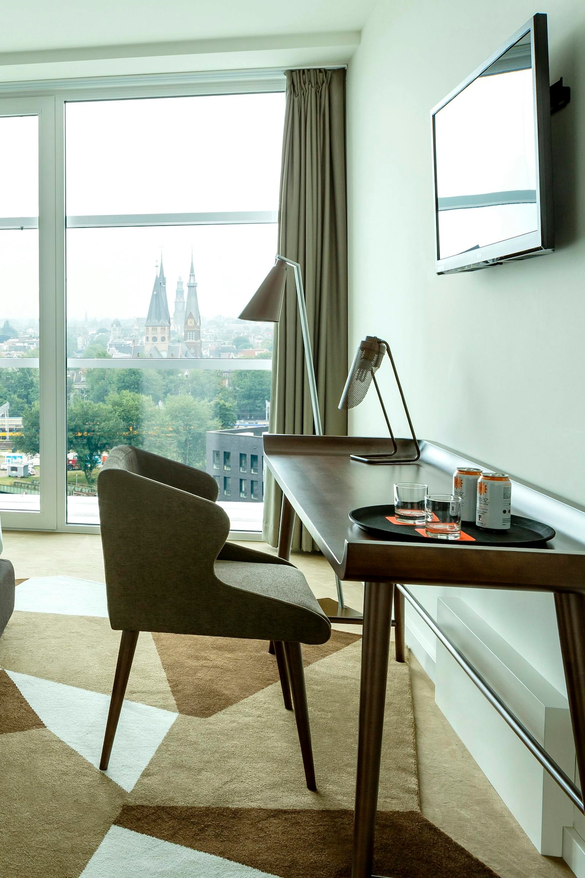 A modern workspace is shown, featuring a wooden desk paired with a comfortable grey chair. A lamp and a flat-screen TV are visible above the desk. Large windows offer a view of a cityscape, including notable spires in the distance, while natural light fills the room.