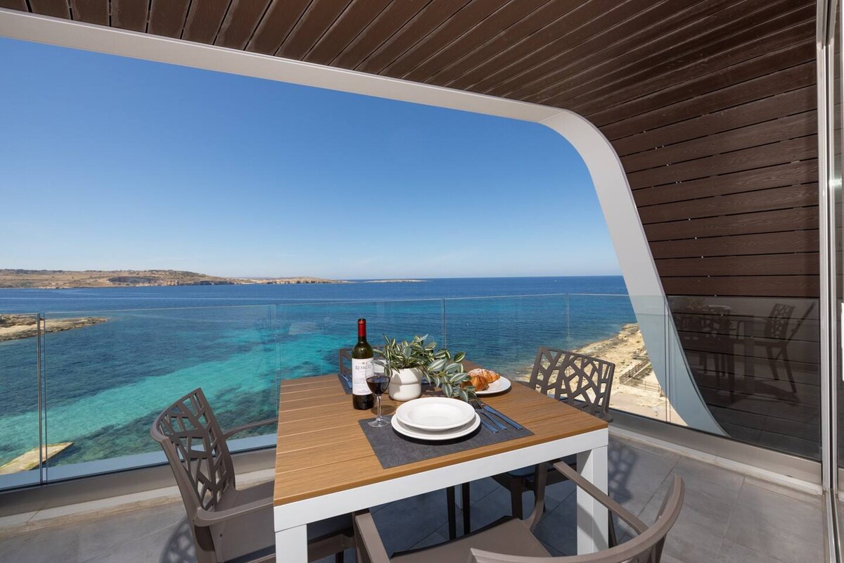 A dining table is set on a balcony with views of the tranquil sea. Several plates and a bottle of wine are arranged atop the table, which is surrounded by grey chairs. The backdrop showcases clear blue water and a sunny sky, enhancing the inviting atmosphere.