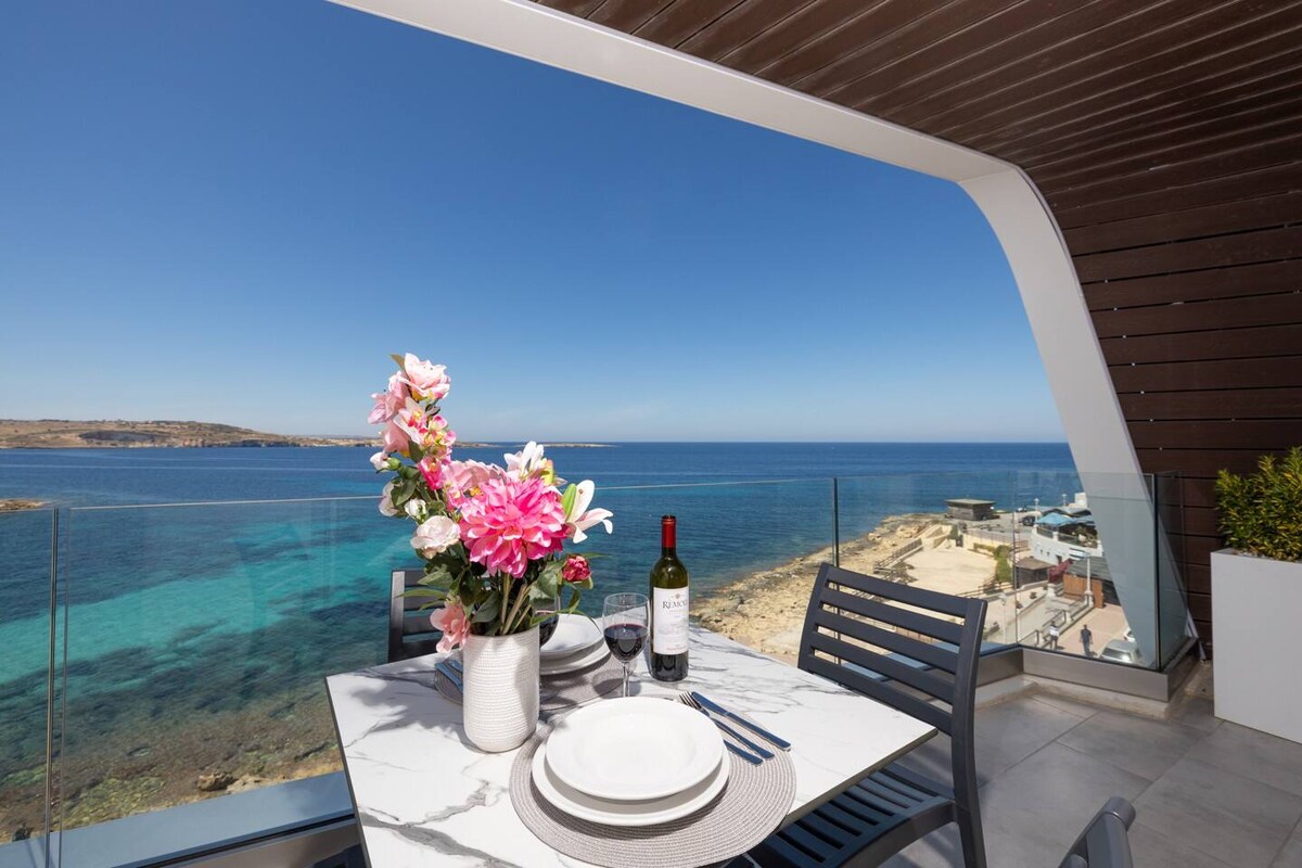 A dining table set for two features elegant tableware, including a vase of pink flowers. The table is positioned on a balcony with a panoramic view of the clear blue waters and coastline, providing an inviting space to enjoy meals outdoors.