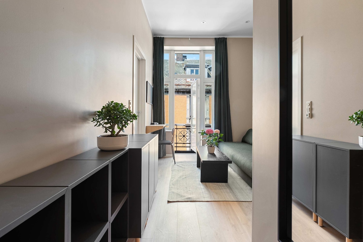 A well-lit living space features a sleek console table with potted plants, contrasting with the warm tones of the walls. Natural light enters through large windows with drapes, highlighting a comfortable sofa and stylish coffee table. A mirror reflects the inviting atmosphere of the area.