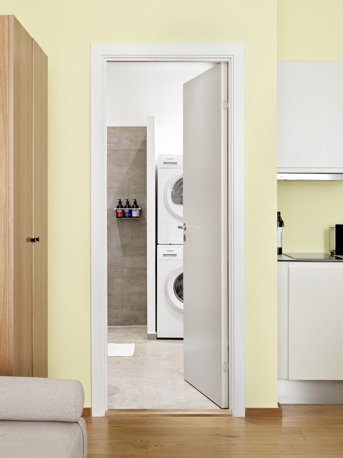 A doorway transitions into a laundry area, where stacked washing machines are visible against a light grey wall. Natural light highlights the smooth, neutral-toned floor. The light yellow walls create a soft backdrop, enhancing the sense of space.