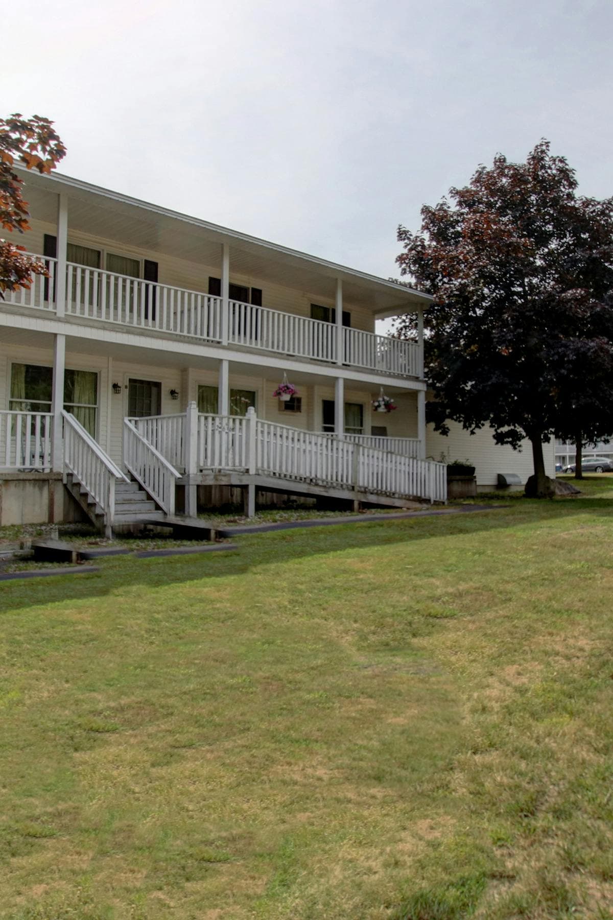 An exterior view of a two-story building features a white railing along the balcony. Steps lead to the entrance, bordered by a landscaped area with green grass. A tree with dark foliage stands nearby, enhancing the outdoor setting.
