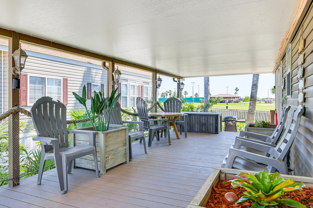 A covered deck features comfortable seating arrangements, including gray Adirondack chairs and side tables. Planters with greenery enhance the space, while a view of the community area can be seen in the background.