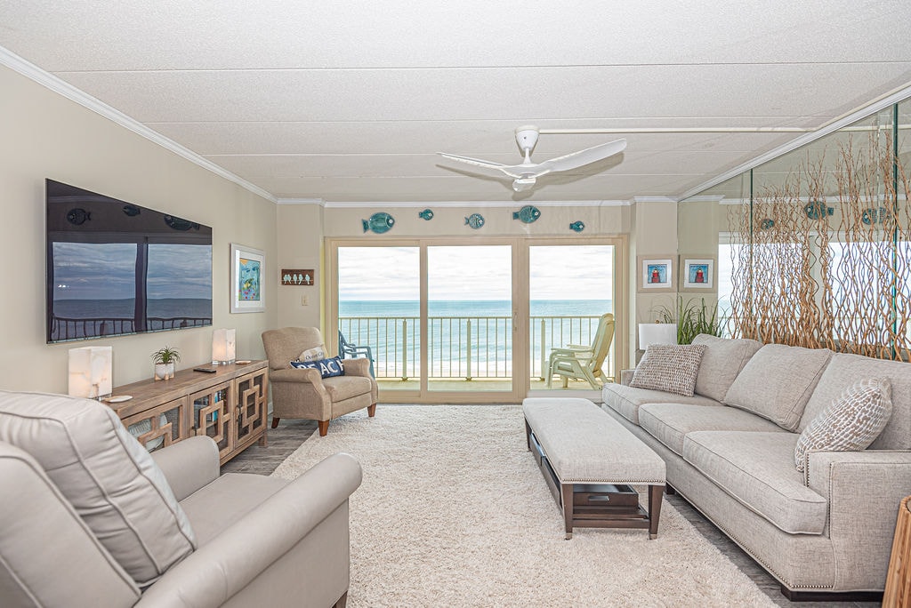 A spacious living area is centered around a large sectional sofa and a cozy armchair. Floor-to-ceiling windows provide uninterrupted views of the ocean. A flat-screen television is mounted on the wall opposite, and plants add a touch of greenery to the room.