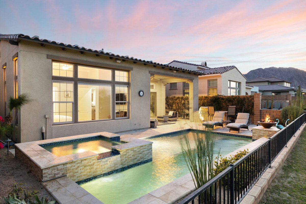 A modern backyard featuring a private pool and hot tub, complemented by stone tiling. Comfortable lounge chairs are arranged around the pool area, with a serene landscape and mountain view in the background, illuminated by a soft sunset glow.