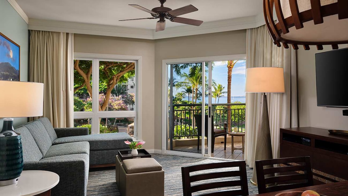 A spacious living area features a comfortable sofa and a stylish coffee table. Large windows frame views of palm trees and distant ocean, allowing natural light to brighten the room. A dining area is visible, equipped with a wooden table and chairs.