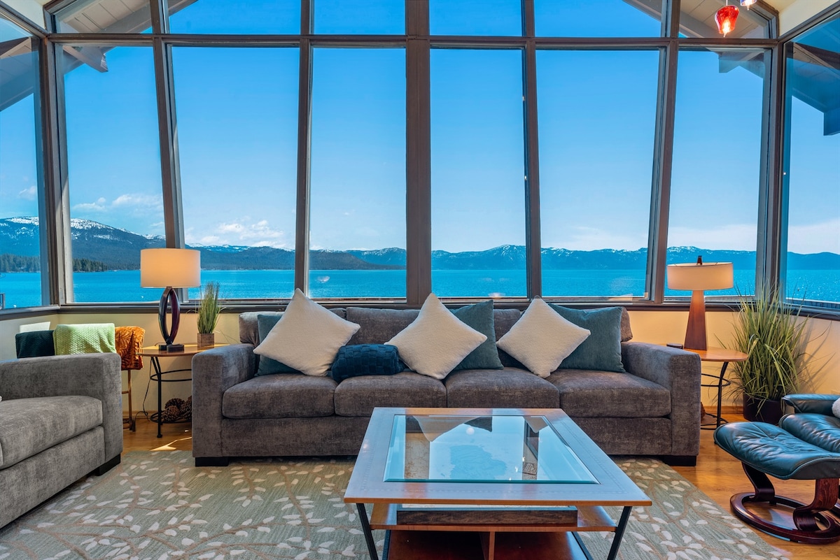 The living room features oversized windows that provide panoramic views of Lake Tahoe. A comfortable sofa with pillows is positioned in front of a glass coffee table. A floor lamp and decorative plants enhance the warm ambiance of the cozy space.