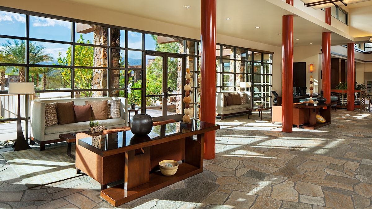 A spacious lobby area features large glass windows allowing natural light to fill the space. Comfortable seating arrangements are positioned around wooden coffee tables. A polished stone floor and natural stone accents blend seamlessly with the surrounding greenery visible through the windows.