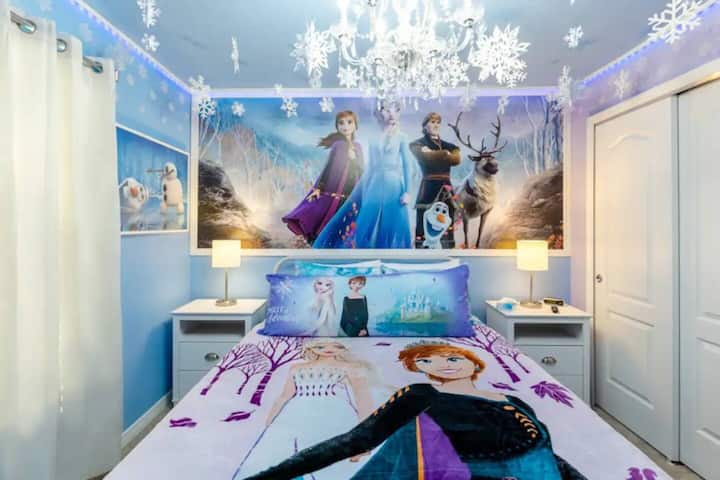 Cama tamaño queen con ropa de cama de Anna y Elsa, calcomanías de copos de nieve en la pared e iluminación azul suave.