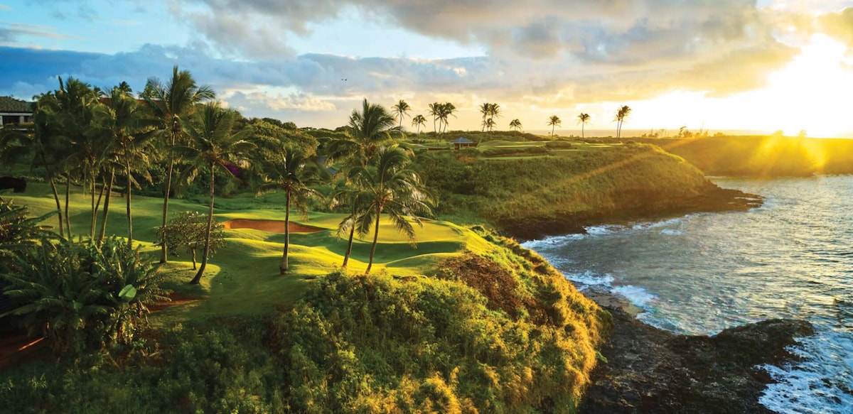 A scenic view of a golf course under a golden sunset, with lush green grass and palm trees lining the coast. Gentle waves crash along the rocky shoreline, enhancing the natural beauty of the landscape.