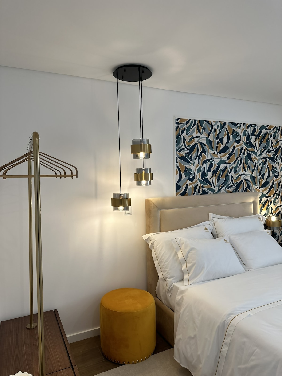 A serene bedroom features a plush bed with white linens and a stylish headboard. Unique pendant lights hang above, illuminating the space. A gold accent stool adds a pop of color next to a wooden nightstand, complementing the vibrant, patterned wall art.