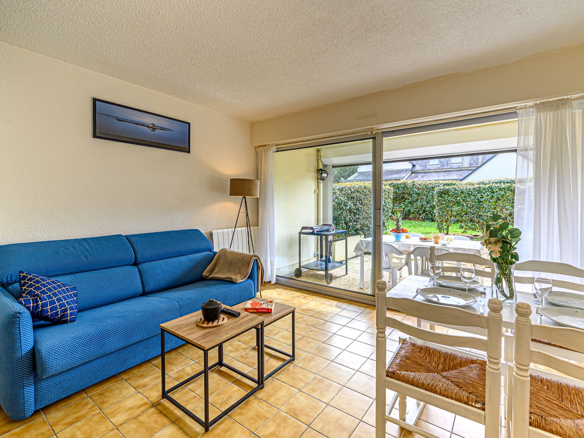 A living area features a blue sofa, a square coffee table, and a dining set for four. Large sliding glass doors allow natural light and offer a view of the terrace area. A floor lamp stands beside the sofa, enhancing the room's comfort.