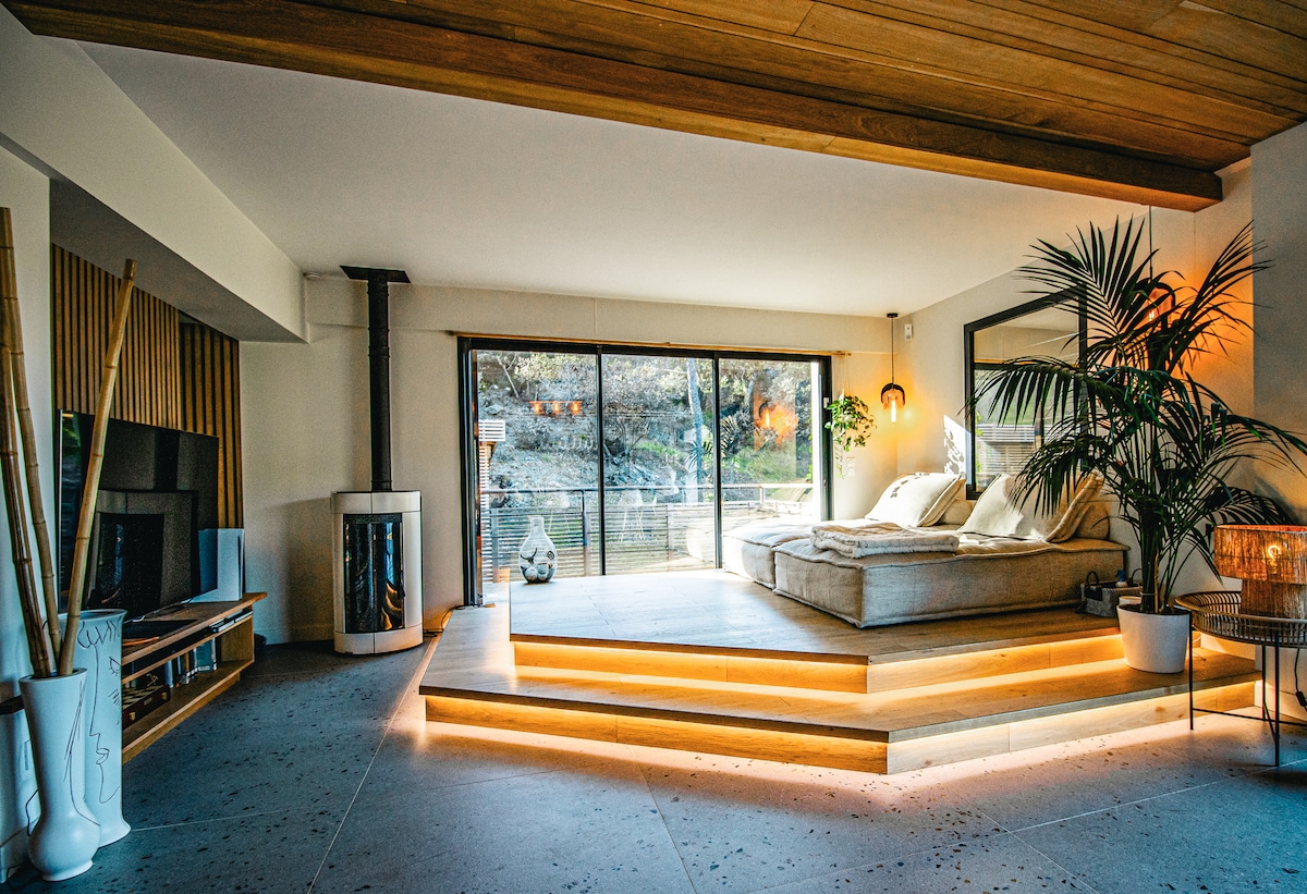 A spacious living area features a low, platform seating arrangement adorned with soft cushions. Natural light floods in through large glass doors, which offer views of the outdoor space. A fireplace and a decorative plant are positioned in the room, adding to the relaxed ambiance.
