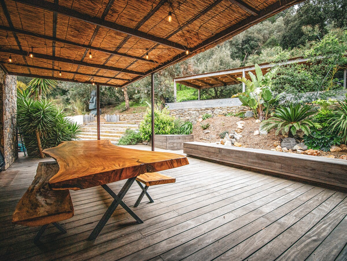 An outdoor dining area features a large wooden table with unique grain patterns, complemented by matching benches. Surrounding greenery includes tropical plants and textured stone, while a wooden deck provides a spacious, natural setting for gatherings. Soft lighting hangs above, enhancing the welcoming ambiance.