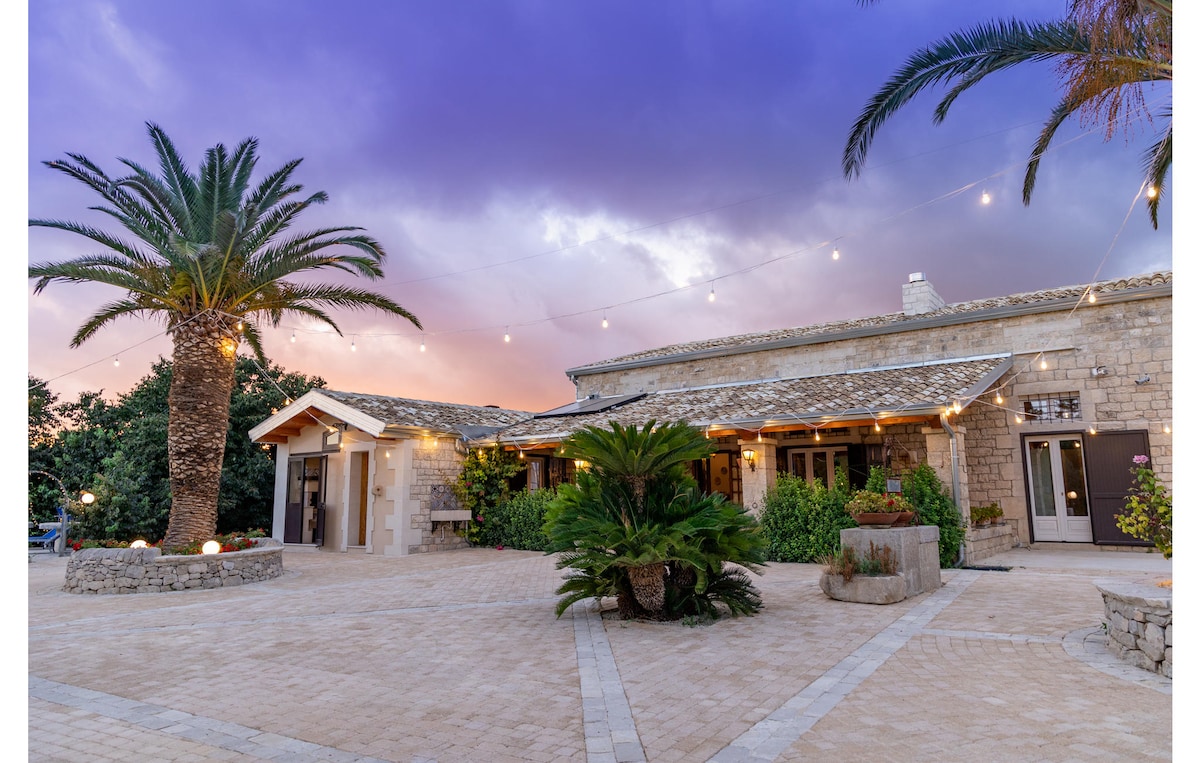 The exterior of the vacation home features a stone façade with large windows, surrounded by palm trees. Soft lighting and a warm sunset create an inviting atmosphere, while landscaped greenery enhances the serene countryside setting.
