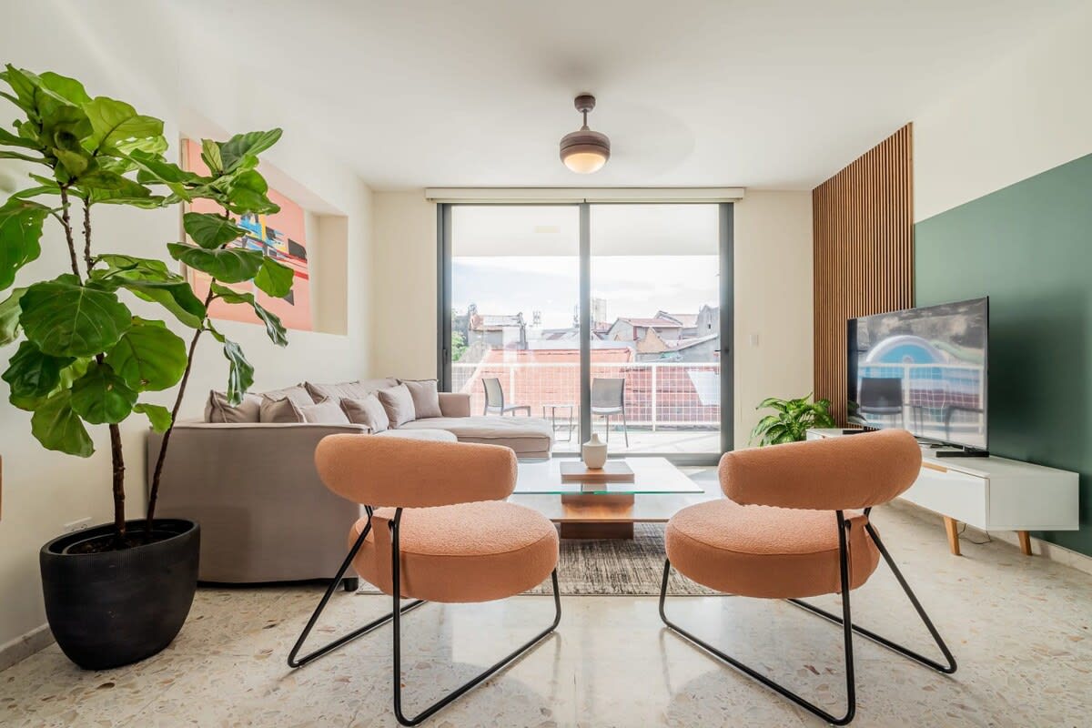 A bright living area is furnished with a comfortable sectional sofa and two stylish chairs. A large glass door allows natural light to fill the space, showcasing a view of the city. A coffee table is centered, complemented by a decorative plant in a pot.