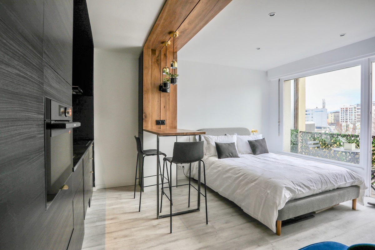 The studio features a modern sleeping area with a spacious bed dressed in white linens, complemented by a stylish wooden accent wall. A small dining table with high chairs is positioned adjacent to the kitchen area. A large glass door provides natural light and access to a furnished balcony.
