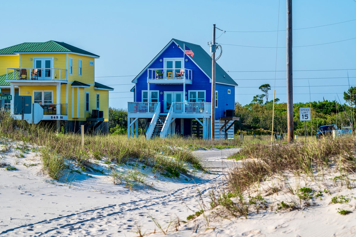 A vibrant blue beach house stands prominently amidst soft sandy dunes, with a yellow house adjacent. The exterior showcases a welcoming entryway with steps leading to a spacious porch adorned with an American flag and colorful outdoor furniture.