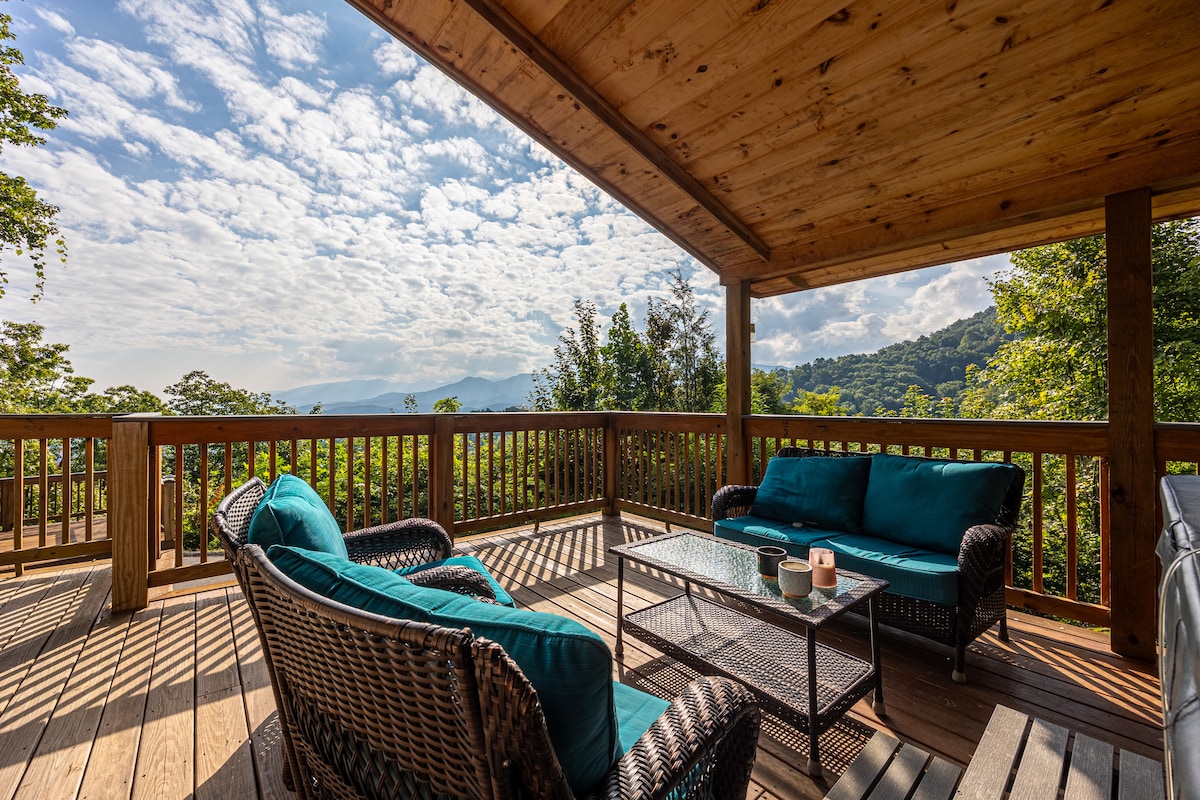 A spacious deck offers a serene seating area with comfortable wicker chairs and a glass-top table. Lush green hills and distant mountains serve as a scenic backdrop, while soft clouds drift across the blue sky, creating an inviting outdoor space to relax.