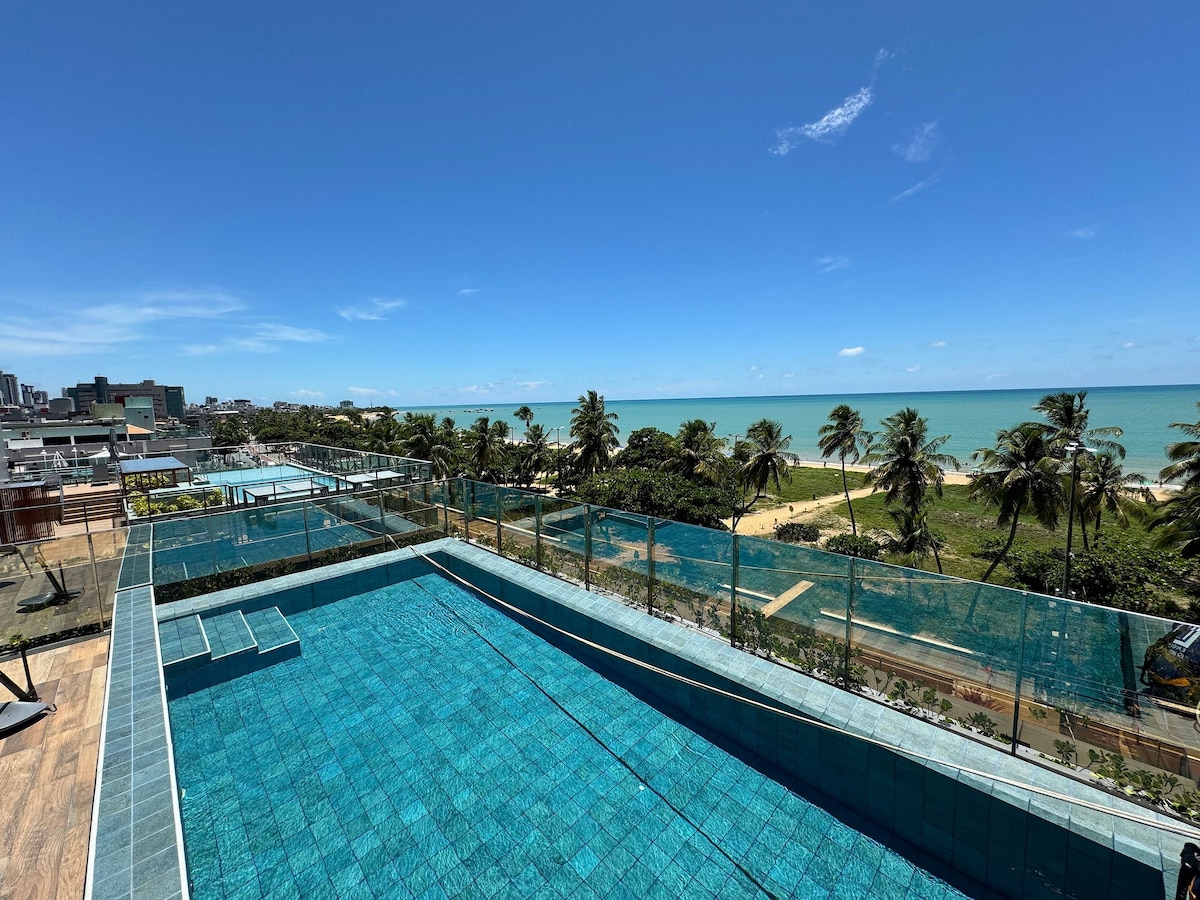An inviting rooftop pool is visible, featuring clear blue water and a stunning view of the ocean. Lush palm trees line the beach, creating a tranquil atmosphere. The pool area is surrounded by glass fencing, enhancing the unobstructed coastal scenery.