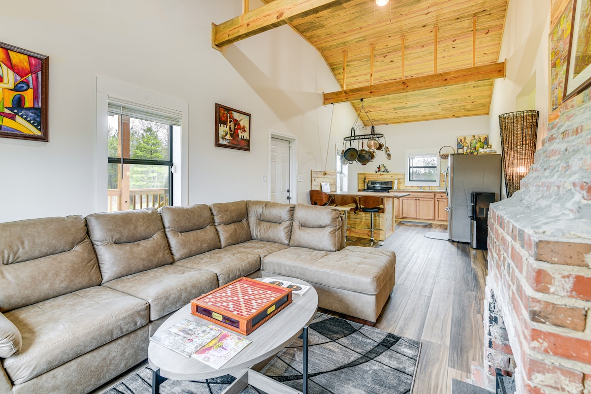An airy living area features a spacious L-shaped sectional sofa, accentuated by a coffee table displaying magazines. A rustic stone fireplace adds character, while wooden beams and a high ceiling enhance the openness. Natural light filters through large windows overlooking the outdoor scenery.