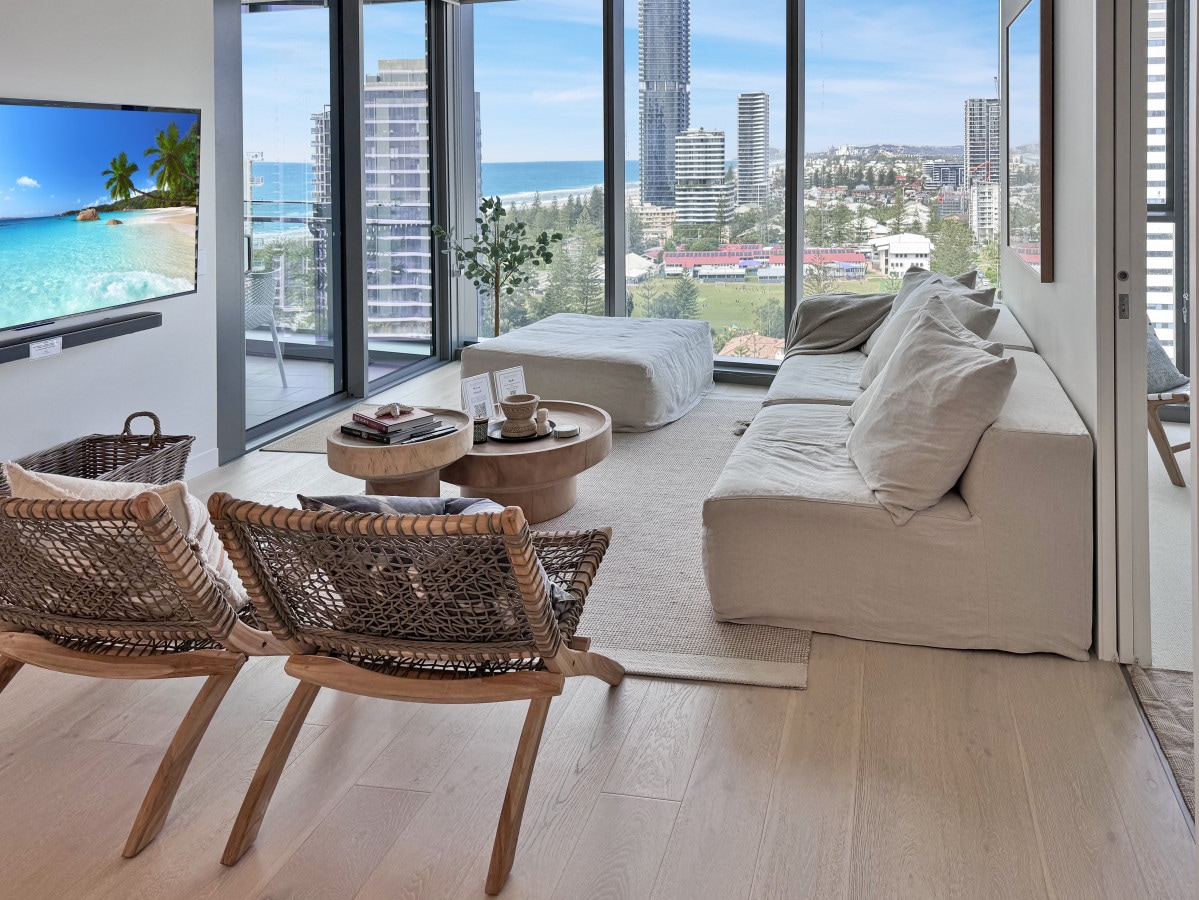 A spacious living area features large floor-to-ceiling windows that frame ocean views. A comfortable sofa and two woven chairs are arranged around a circular coffee table. Natural light fills the room, highlighting the light-colored flooring and contemporary decor.