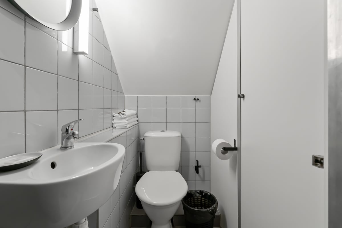 A compact bathroom features white tiled walls and a sloped ceiling. A modern sink is positioned beside a toilet, with neatly stacked towels displayed nearby. A wastebasket is located on the floor, and a toilet paper holder is mounted on the wall.